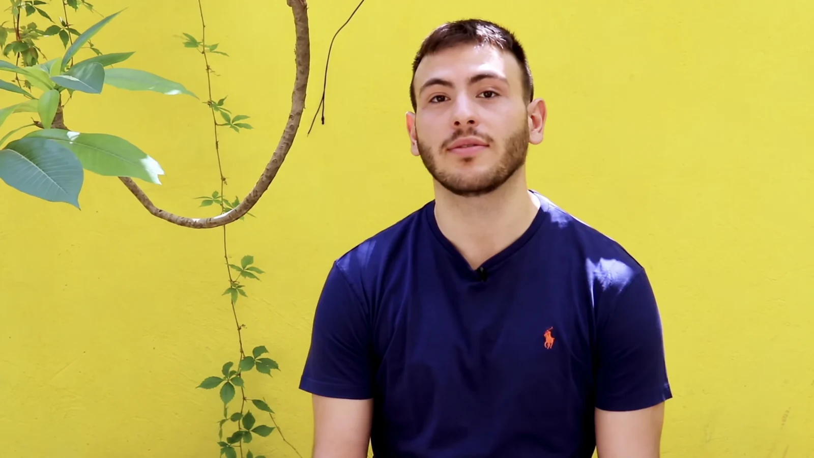 Alex speaking in his ActingLab interview, against a yellow wall and climbing greenery.