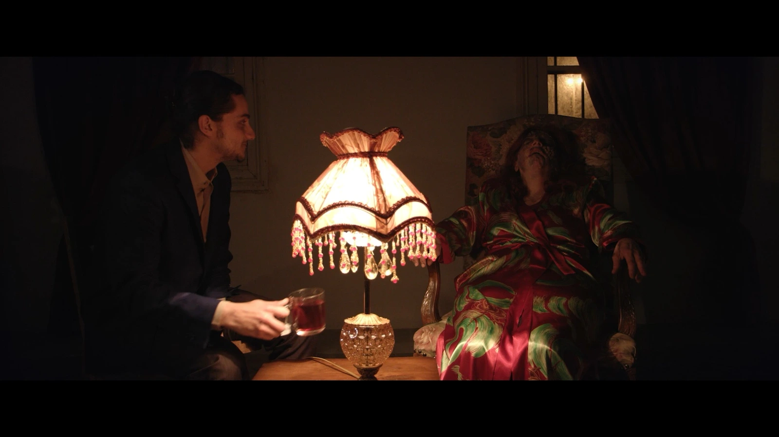 Two figures seated beside a glowing fringed table lamp in a warm, richly furnished interior. · Stitched up Bonds