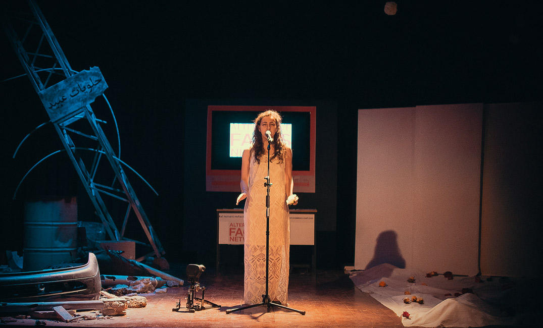 A vocalist at a microphone under a warm spotlight, beside blue-lit scenery and rubble.